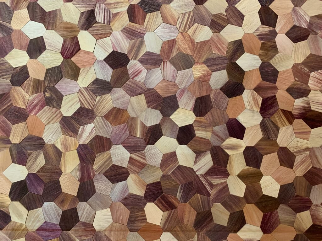 a close-up view of a mosaic pattern made from various pieces of wood, each with distinct textures and shades. The wood pieces are arranged in a geometric pattern, primarily pentagonal shapes, showcasing a variety of natural wood colors ranging from light beige to deep brown and reddish hues. The material properties of the wood pieces include varying grain patterns, textures, and finishes, some appearing smooth and polished while others are more coarse and rugged. The intricate pattern and diverse textures create a visually appealing and organic composition, highlighting the natural beauty and unique characteristics of each wood type.